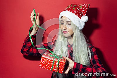 Studio portrait of young modern blonde girl, wearing Santa hat with snowflakes, trying to open a Christmas present with green bow, Stock Photo