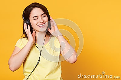 Studio shot of adorable woman with dark hair enjoys of listening to music in headphones, keeps hands on earphones, stands with Stock Photo