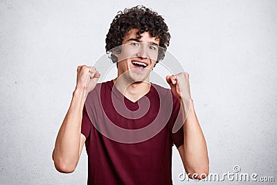 Studio shot of cheerful attractive European male being excited and glad to achieve victory, expresses positivity, clenches fists, Stock Photo