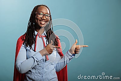 Studio shot of cheerful and optimistic justice defender woman wearing red cape while pointing fingers to right Stock Photo