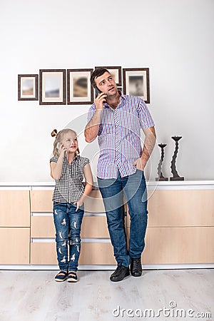 Studio shot of a little girl and her dad standing at home and talking on the phone the same way. Daughter imitates her father Stock Photo