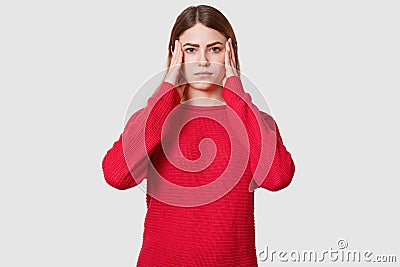 Studio shot of displeased female suffers from headache, dressed in red sweater, keeps hands on temples, has upset facial Stock Photo