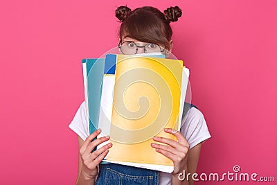 Studio shot of emotive european woman hides behind blue and yellow paper folders, wearing round glasses, white t shirt, denim Stock Photo