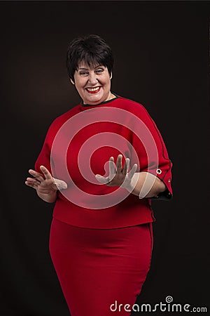 Studio shot of a senior woman wearing a red dress on a dark background. She is smiling cheerfully gesticulating Stock Photo