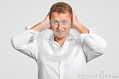 Studio shot of successful young male entrepreneur keeps hands on head, looks seriously at camera, dressed elegantly, being tired o Stock Photo