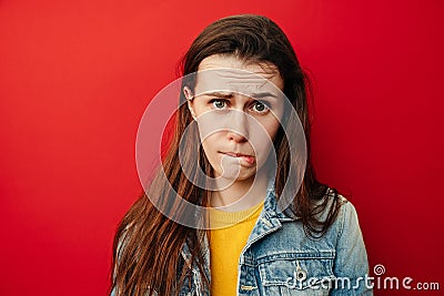 Studio shot of upset young woman purses lower lip, pouting and has displeased look, looks sadly at camera, wears denim jacket, Stock Photo