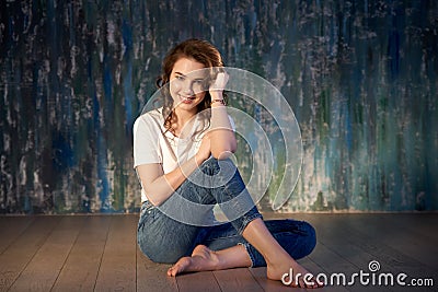 Studio shot of a young smiling girl in jeans and a t-shirt sitting on the floor. Bright sunlight, positive emotions Stock Photo