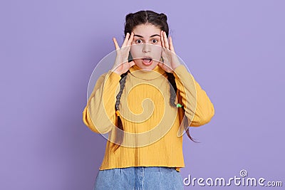 Stunned dark haired woman with two pigtails, keeping hands near face, afraids of something astonishing, dresses in fashionable Stock Photo