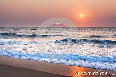 Stunning sunset or sunrise over the sea or ocean on the beach, purple sky, blue waves, white foam and golden sun reflection Stock Photo