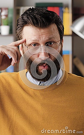 Stupid. Irritated bearded man in glasses in office or apartment room looks at camera and twirls his finger at his temple Stock Photo