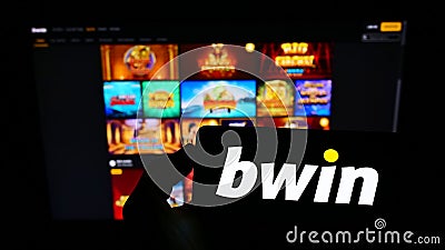 Person holding cellphone with logo of Austrian company bwin Interactive Entertainment AG on screen in front of webpage. Editorial Stock Photo