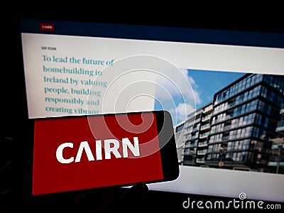 Person holding cellphone with logo of Irish housebuilding company Cairn Homes plc on screen in front of business webpage. Editorial Stock Photo