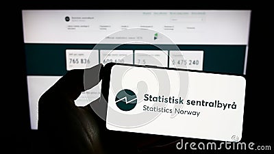 Person holding cellphone with logo of Norwegian agency Statistics Norway (SSB) on screen in front of webpage. Editorial Stock Photo