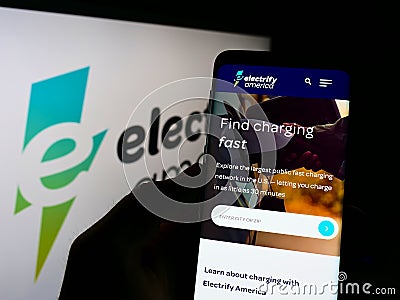 Person holding cellphone with website of US EV charging company Electrify America LLC on screen in front of logo. Editorial Stock Photo