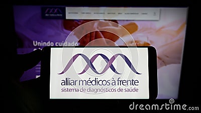 Person holding mobile phone with logo of Brazilian healthcare company Grupo Alliar on screen in front of business web page. Editorial Stock Photo