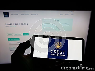 Person holding mobile phone with logo of British housebuilding company Crest Nicholson plc on screen in front of web page. Editorial Stock Photo