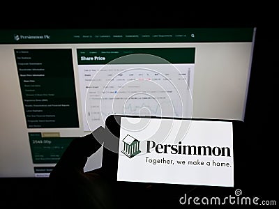 Person holding mobile phone with logo of British housebuilding company Persimmon plc on screen in front of web page. Editorial Stock Photo