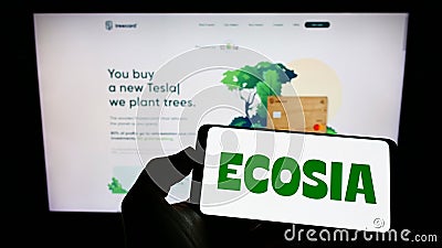 Person holding mobile phone with logo of German search engine company Ecosia GmbH on screen in front of business web page. Editorial Stock Photo