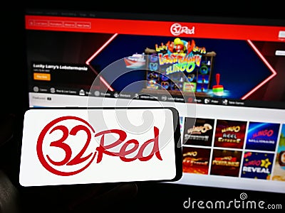 Person holding mobile phone with logo of online casino company 32Red Limited on screen in front of business web page. Editorial Stock Photo