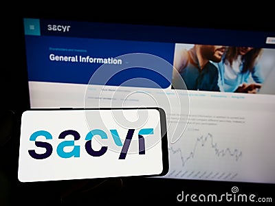 Person holding mobile phone with logo of Spanish infrastructure company Sacyr SA on screen in front of business web page. Editorial Stock Photo