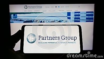 Person holding mobile phone with logo of Swiss investment company Partners Group Holding AG on screen in front of web page. Editorial Stock Photo