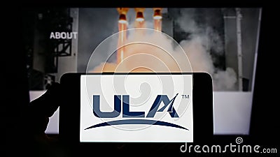 Person holding mobile phone with logo of US space company United Launch Alliance (ULA) on screen in front of web page. Editorial Stock Photo