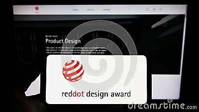 Person holding smartphone with logo of German international prize Red Dot Design Award on screen in front of website. Editorial Stock Photo