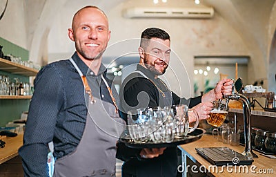 Stylish bearded barman dressed black uniform beer tapping at bar counter and waiter with tray chatting smiling to each other. Stock Photo