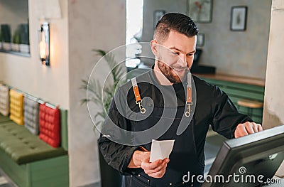 Stylish bearded smiling waiter dressed black uniform processing customer orders using point of sale order terminal system touch Stock Photo