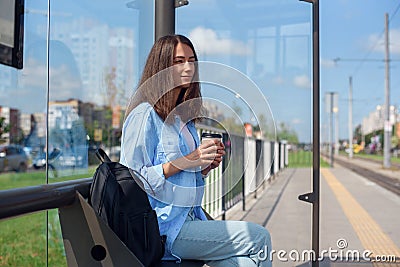 Beautiful girl waits for subway or tram on public transport station in the morning. Happy young woman with cup of coffee Stock Photo