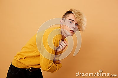 Stylish blond guy dressed in yellow sweater and black jeans is posing on the beige background in the studio Stock Photo