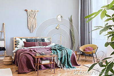 Stylish boho interior of bedroom with design furnitures, honey yellow armchair, macrame,lamp, plant and elegant accessories. Stock Photo