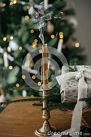 Stylish candle, wrapped christmas gift, rustic basket with fir branches against festive decorated tree in scandinavian room. Moody Stock Photo