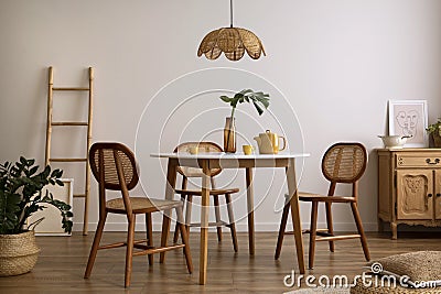 The stylish dining room with round table, rattan chair, wooden commode, pock up poster and kitchen accessories. Beige wall with Stock Photo