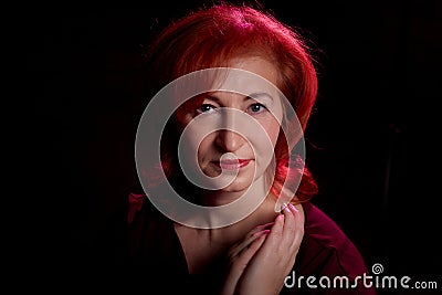 Stylish elderly woman with red hair in vinous dress posing in studio with dark background and red flash and light Stock Photo