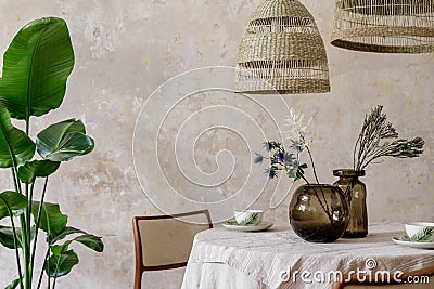 Stylish and elegant dining room interior with diner table, design chairs, rattan pendant lamps, cups, bouquet of dried flowers. Stock Photo