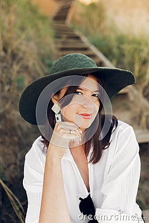 Stylish girl in fashionable look sitting on wooden stairs on beach cliff at sea in sunny light. Happy young boho woman in hat Stock Photo