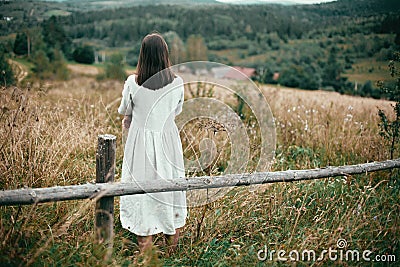 Stylish girl in linen dress standing at aged wooden fence among herbs and wildflowers, looking at field. Boho woman relaxing in Stock Photo