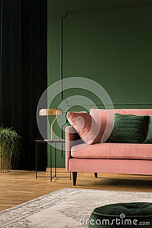 Stylish golden lamp on industrial table next to pink couch with emerald pillows in dark green living room interior,copy space on Stock Photo