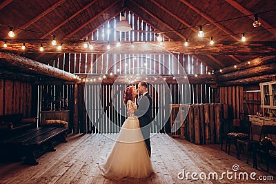 Stylish groom and happy bride hugging under retro bulbs lights in wooden barn. rustic wedding concept, space for text. newlyweds Stock Photo