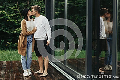 Stylish hipster couple hugging and kissing on wooden terrace, relaxing in cabin in mountains. Happy young family in modern outfits Stock Photo