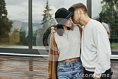 Stylish hipster couple hugging on wooden porch, relaxing in modern cabin in mountains. Happy young family in modern outfits Stock Photo