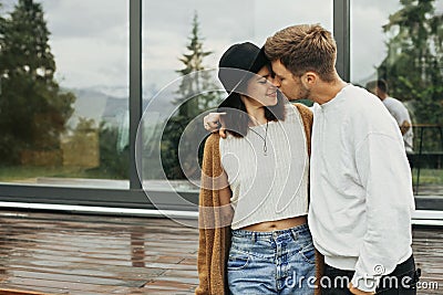 Stylish hipster couple hugging on wooden porch, relaxing in modern cabin in mountains. Happy young family in modern outfits Stock Photo