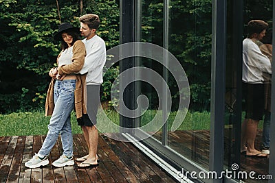 Stylish hipster couple hugging on wooden terrace, relaxing in cabin in mountains. Happy young family in modern outfits embracing Stock Photo