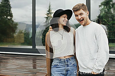 Stylish hipster couple smiling on wooden porch, relaxing in modern cabin in mountains. Happy young family in modern outfits Stock Photo