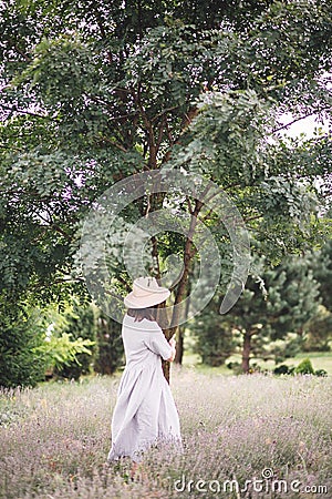 Stylish hipster girl in linen dress and hat relaxing in lavender field near tree. Happy bohemian woman enjoying summer vacation in Stock Photo