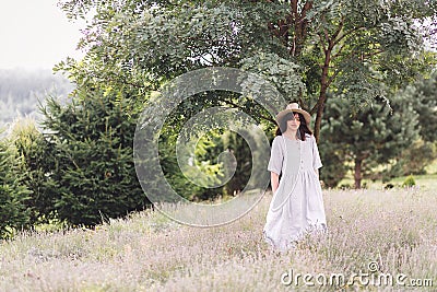 Stylish hipster girl in linen dress and hat relaxing in lavender field near tree. Happy bohemian woman enjoying summer vacation in Stock Photo