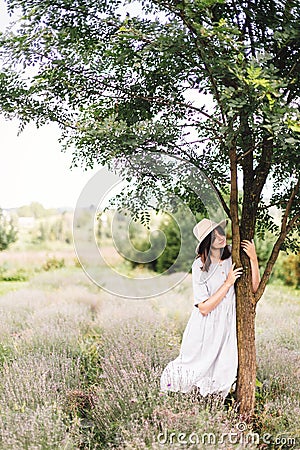 Stylish hipster girl in linen dress and hat relaxing in lavender field near tree. Happy bohemian woman enjoying summer vacation in Stock Photo