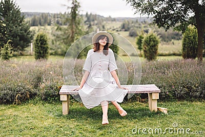 Stylish hipster girl in linen dress and hat sitting on bench at lavender field and relaxing in the morning. Happy bohemian woman Stock Photo