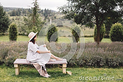 Stylish hipster girl in linen dress and hat sitting on bench at lavender field and relaxing in the morning. Happy bohemian woman Stock Photo